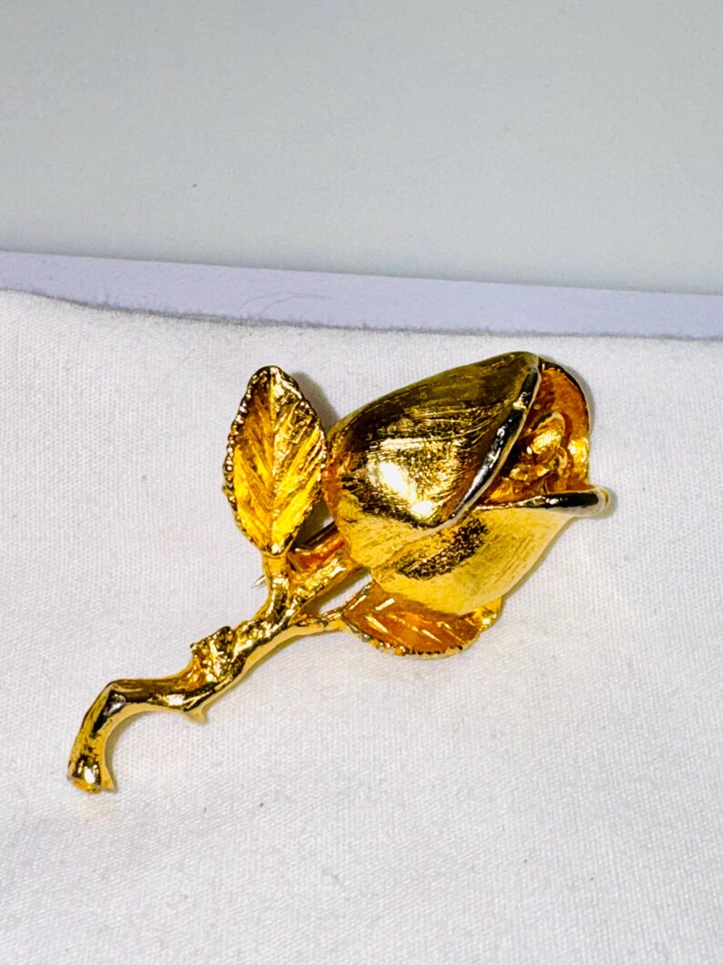 K&ouml;nnte beinhalten: Eine goldfarbene Metallbrosche in Form einer Rosenknospe an einem Stiel mit einem einzelnen Blatt. Die Oberfl&auml;che der Blume und des Stiels hat eine texturierte, reflektierende Oberfl&auml;che, die das Licht auf den geschwungenen Bl&uuml;tenbl&auml;ttern einf&auml;ngt.