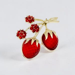 May include: A gold-tone brooch shaped like two strawberries with red enamel bodies and textured surfaces. The piece features three small stems topped with clusters of sparkling red crystals and gold-colored leaves.