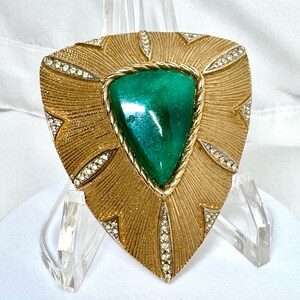May include: A triangular gold-tone brooch featuring a central smooth green stone framed by a twisted rope border. The textured gold surface radiates outward, accented by six pointed inlays set with small clear crystals along the edges.