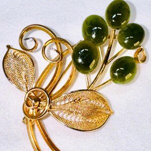 May include: A gold-tone metal brooch featuring two filigree leaves and swirling wire stems. Attached to the stems are four smooth, round, dark olive-green cabochon stones arranged in a cluster, resembling small berries or fruit on a branch.