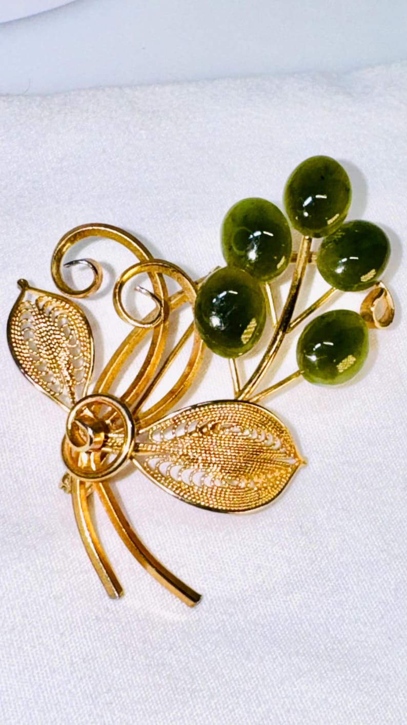 May include: A gold-tone metal brooch featuring two filigree leaves and swirling wire stems. Attached to the stems are four smooth, round, dark olive-green cabochon stones arranged in a cluster, resembling small berries or fruit on a branch.