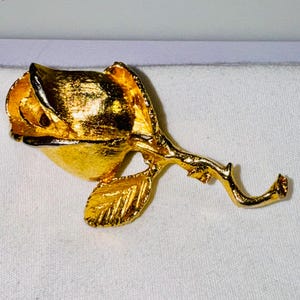K&ouml;nnte beinhalten: Eine Brosche aus goldfarbenem Metall in Form einer Rosenknospe. Das St&uuml;ck verf&uuml;gt &uuml;ber eine texturierte, geb&uuml;rstete Goldoberfl&auml;che auf den Bl&uuml;tenbl&auml;ttern und ein einzelnes Blatt an einem geschwungenen Stiel. Das Design zeigt eine geschlossene Bl&uuml;te.
