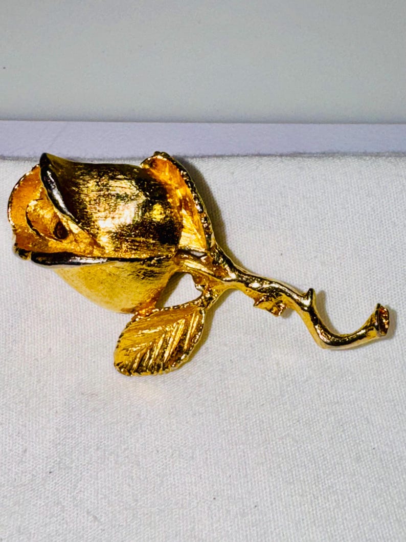 K&ouml;nnte beinhalten: Eine Brosche aus goldfarbenem Metall in Form einer Rosenknospe. Das St&uuml;ck verf&uuml;gt &uuml;ber eine texturierte, geb&uuml;rstete Goldoberfl&auml;che auf den Bl&uuml;tenbl&auml;ttern und ein einzelnes Blatt an einem geschwungenen Stiel. Das Design zeigt eine geschlossene Bl&uuml;te.