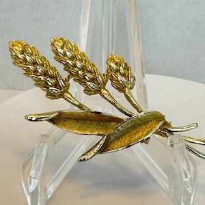 Könnte beinhalten: Eine goldfarbene Brosche mit einem Weizenährendesign. Die Brosche hat drei Weizenähren und zwei Blätter, alle mit detaillierten Texturen. Die Brosche wird auf einem transparenten Ständer vor neutralem Hintergrund präsentiert.