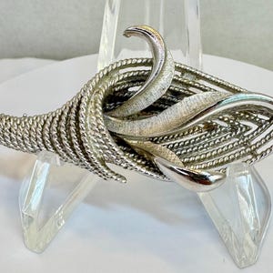May include: A silver-toned brooch with a textured, openwork design. The brooch features a swirling, leaf-like shape with a braided rope texture and polished accents. A stylish accessory.