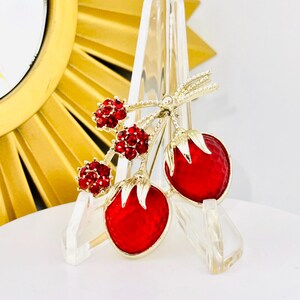 May include: A gold-tone brooch featuring two red enameled strawberries with textured surfaces and three clusters of small, round, red rhinestones attached to a stem with gold leaves. The brooch has a polished finish and intricate detailing.