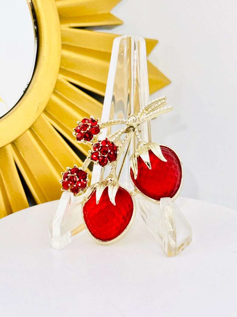 May include: A gold-tone brooch featuring two red enameled strawberries with textured surfaces and three clusters of small, round, red rhinestones attached to a stem with gold leaves. The brooch has a polished finish and intricate detailing.