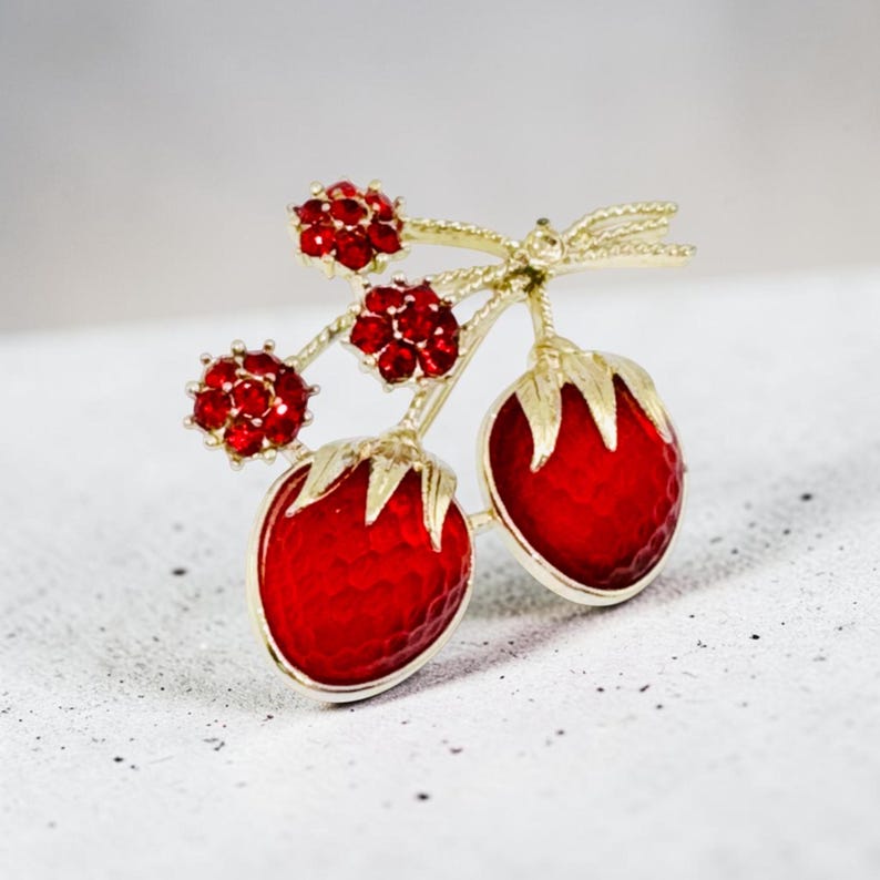 May include: A gold-tone brooch shaped like two strawberries with textured red enamel surfaces and green leaves. The design includes three small stems topped with clusters of sparkling red rhinestones, creating a detailed fruit-inspired accessory.