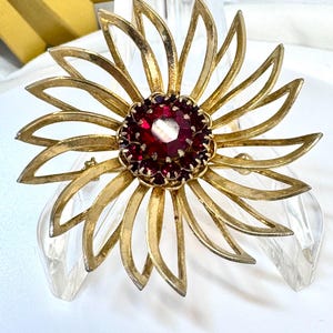 May include: A gold-tone metal brooch shaped like a flower with elongated, pointed petals. The center features a large, round, faceted deep red stone surrounded by a ring of smaller, matching red stones set in a circular metal frame.