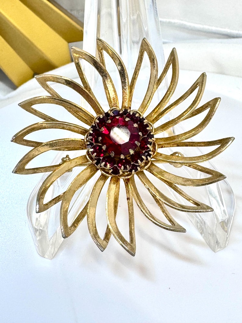 Op de afbeelding: Een goudkleurige metalen broche in de vorm van een bloem met langwerpige, puntige bloemblaadjes. Het midden bevat een grote, ronde, gefacetteerde donkerrode steen omringd door een ring van kleinere, bijpassende rode steentjes in een metalen zetting.