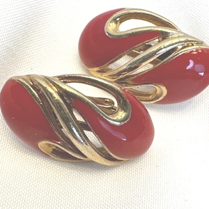 May include: A pair of vintage clip-on earrings. Each earring features a glossy red oval shape with a gold-tone metal swirl design. The earrings are set against a white background.