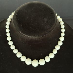 May include: A cream-coloured beaded necklace displayed on a black velvet bust. The necklace features graduated round beads, with the largest beads at the centre and smaller beads towards the clasp. The beads have a smooth, glossy finish.