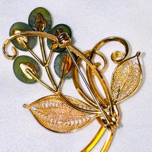 May include: A gold-tone brooch featuring a floral design with four round, dark green stone accents and two filigree-style leaves. The back of the brooch displays a metal pin mechanism with a rotating clasp, showing the reverse side of the piece.