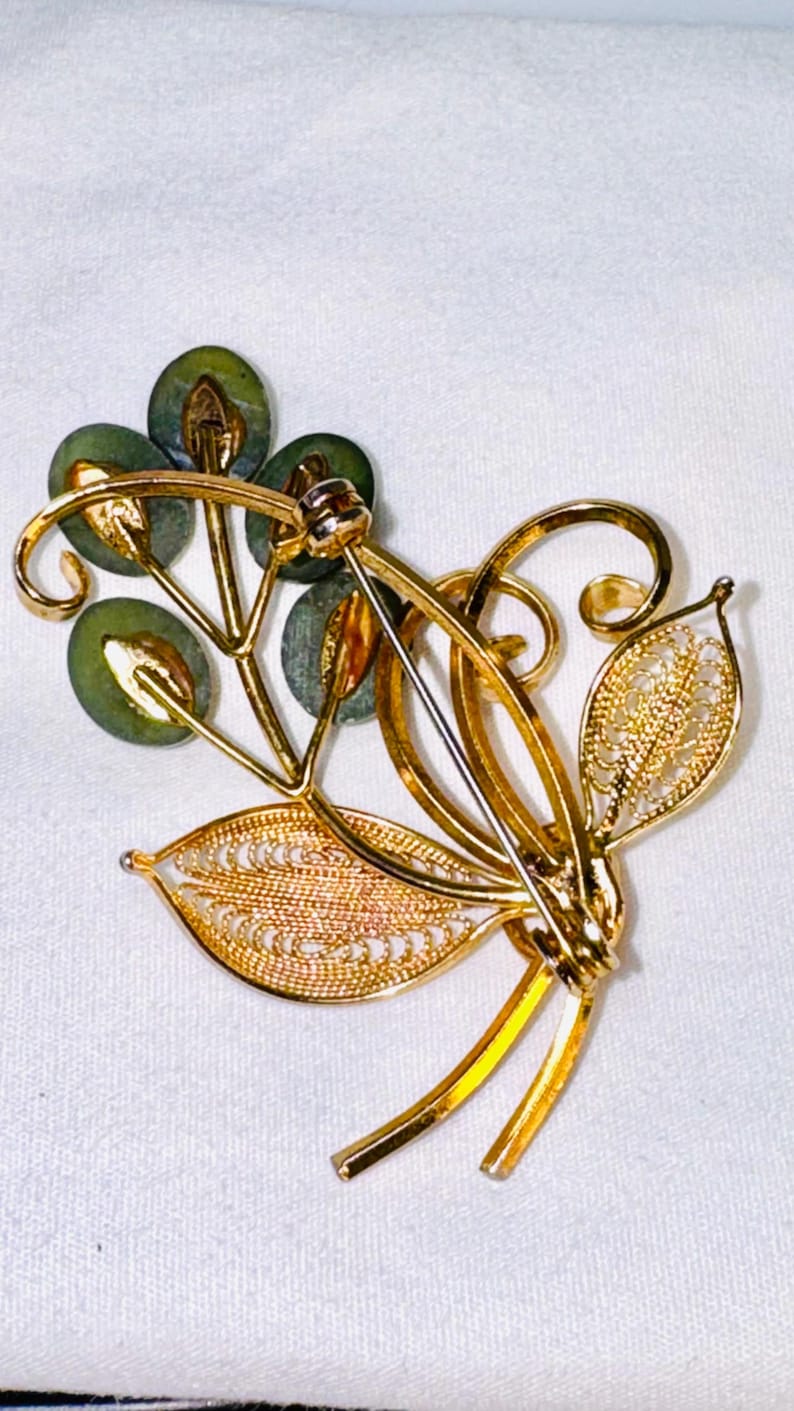 May include: A gold-tone brooch featuring a floral design with four round, dark green stone accents and two filigree-style leaves. The back of the brooch displays a metal pin mechanism with a rotating clasp, showing the reverse side of the piece.