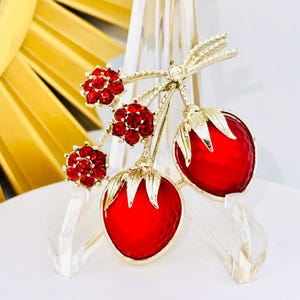 May include: A gold-tone brooch featuring two red strawberry-shaped charms with textured surfaces and green metal leaves, accented by three small clusters of round red rhinestones attached to thin gold-tone stems and a small clear stone.