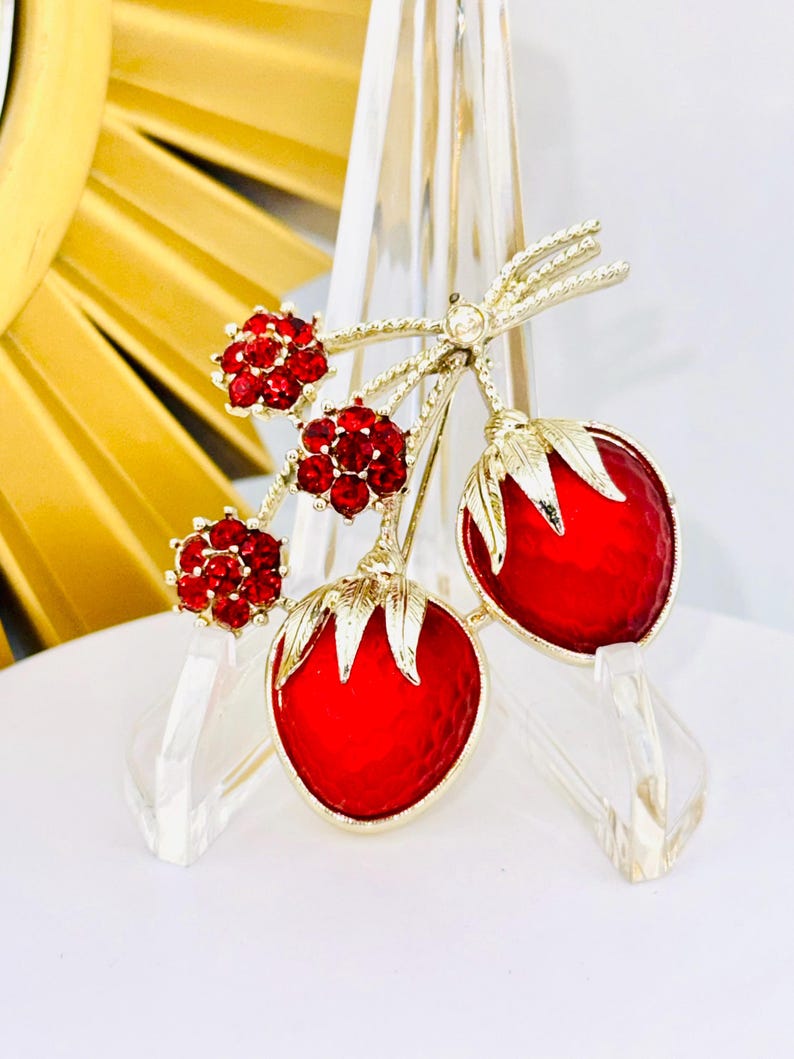 May include: A gold-tone brooch featuring two red strawberry-shaped charms with textured surfaces and green metal leaves, accented by three small clusters of round red rhinestones attached to thin gold-tone stems and a small clear stone.