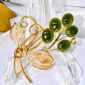 May include: A gold-toned brooch featuring two delicate filigree leaves and swirling wire stems. Attached to the stems are four smooth, oval-shaped dark green stones that resemble small berries or olives, creating a botanical-inspired design.