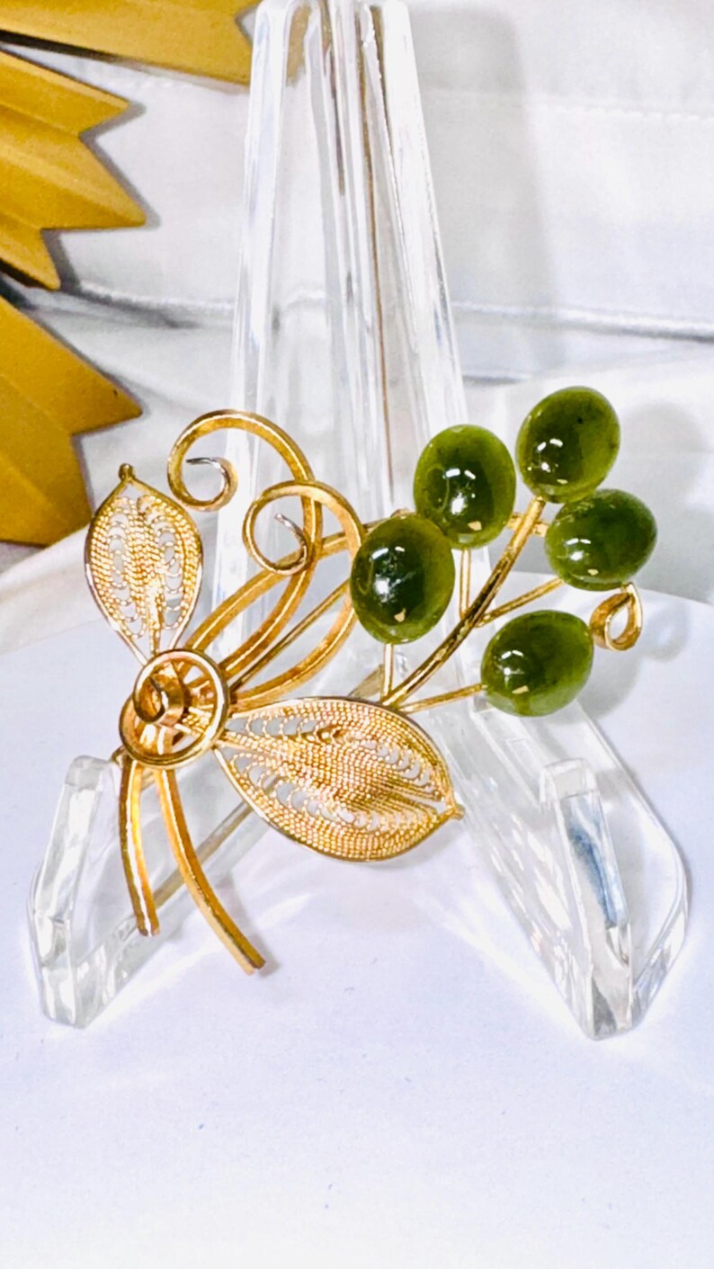May include: A gold-toned brooch featuring two delicate filigree leaves and swirling wire stems. Attached to the stems are four smooth, oval-shaped dark green stones that resemble small berries or olives, creating a botanical-inspired design.