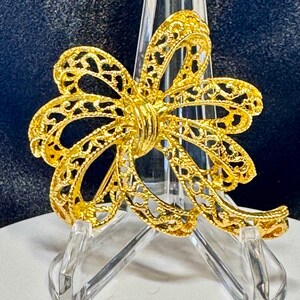 May include: A gold-colored bow brooch with intricate openwork detailing. The brooch has a classic bow shape with multiple loops and a central knot. The metal has a shiny finish, and the brooch is displayed on a clear stand.