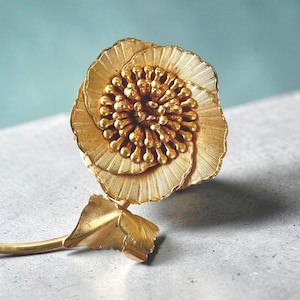 May include: A gold-colored, flower-shaped brooch with detailed petal and center designs. The brooch has a stem and leaf detail, set against a light blue and gray background. The brooch is likely made of metal.