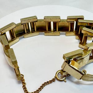 May include: A gold-toned bracelet with a rectangular link design. The bracelet features a safety chain and a clasp closure. The links are connected by vertical bars, creating an open, airy feel. The bracelet is displayed on a white surface.