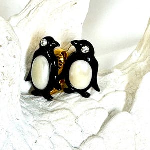 Könnte beinhalten: Ein Paar Ohrringe in Pinguinform. Jeder Ohrring hat einen schwarzen Körper, einen weißen Bauch und einen kleinen, funkelnden Edelstein auf dem Kopf. Die Ohrringe sind auf goldfarbenen Stiften befestigt.