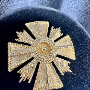 Könnte beinhalten: Goldfarbene Kreuzbrosche mit strukturiertem Design. Die Mitte hat ein kreisförmiges Element mit einem kleinen, erhabenen Detail. Die Kreuzarme haben ein strukturiertes, fächerartiges Muster und sind von einer gedrehten, seilartigen Bordüre umrandet.