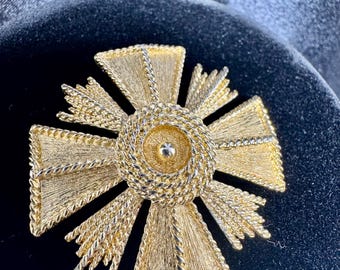 Op de afbeelding: Goudkleurige kruisvormige broche met een textuurontwerp. Het midden heeft een cirkelvormig element met een klein, verhoogd detail. De kruisarmen hebben een textuur, waaiervormig patroon en zijn omzoomd door een gedraaide, touwachtige rand.