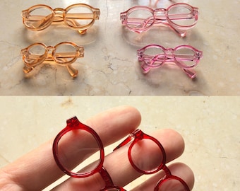 May include: A collection of miniature toy glasses in various colors including red, orange, and pink. The frames are round and made of a translucent plastic material. Some pairs are larger than others, featuring thin temples and clear lenses.