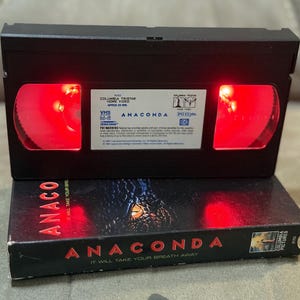 May include: A black VHS tape labeled "Anaconda" with red glowing reels. The VHS case is also black with the movie title in red. The text "It Will Take Your Breath Away" is also visible.