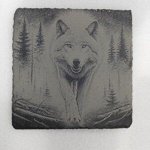 May include: A square, gray slate coaster featuring an engraved image of a wolf. The wolf is depicted in a forest setting with pine trees and a dark background. The wolf is facing forward with its mouth slightly open.