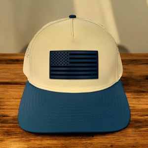 May include: A baseball cap with a cream-colored crown and a blue bill. The front of the cap features a dark blue American flag patch. The cap has a curved bill and a button on top.