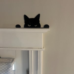 May include: Black silhouette of a cat peeking over a white door frame. The cat has a grumpy expression.