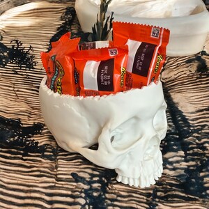 May include: A white skull-shaped container filled with orange Reese's peanut butter cup candies. The container has a detailed, textured surface and is placed on a patterned surface. The candies have the text "Reese's" and expiration dates printed on the wrappers.