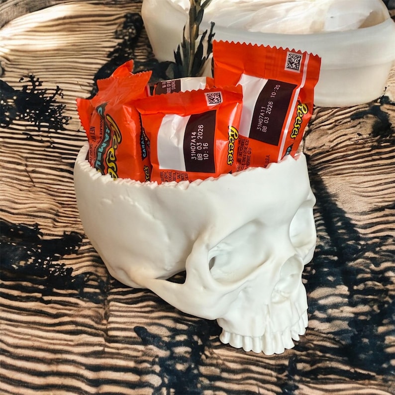 May include: A white skull-shaped container filled with orange Reese's peanut butter cup candies. The container has a detailed, textured surface and is placed on a patterned surface. The candies have the text "Reese's" and expiration dates printed on the wrappers.