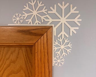 May include: White snowflake wall decor in various sizes, arranged in a corner on a light blue wall. The snowflakes are placed near a wooden door frame. The decor adds a winter or holiday theme to the room.