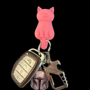 May include: A pink cat-shaped keychain holder with a silver keyring holding a car key fob, a Mandalorian helmet charm, and a multi-tool. The background is black.