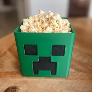 May include: Green popcorn container shaped like a Minecraft Creeper with black eyes and mouth.