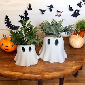 May include: Two white ghost-shaped planters with green plants inside sit on a wooden table. A carved orange pumpkin and a small gourd are nearby. Black bat and tree cutouts decorate the background, creating a Halloween theme.