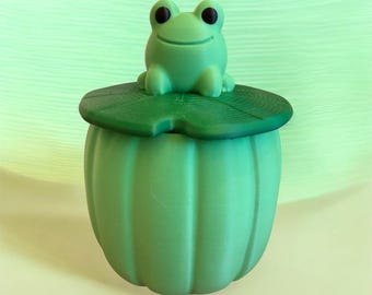 Può includere: Un contenitore verde chiaro con una texture a coste verticali e un coperchio verde scuro a forma di foglia di ninfea. Una piccola figura di rana verde chiaro con occhi neri siede sopra il coperchio, fungendo da maniglia per la chiusura.