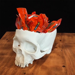 May include: A white skull-shaped bowl filled with orange candy wrappers. The bowl is made of a smooth, matte material and sits on a wooden surface. The candy wrappers are filled with Reese's Caramel Cups.