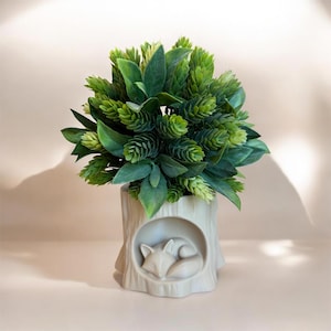 May include: A decorative artificial plant arrangement. The green foliage sits in a cream-colored ceramic pot shaped like a tree stump. A sleeping fox is sculpted into the front of the pot. The plant has a variety of green leaves and cone-shaped buds.
