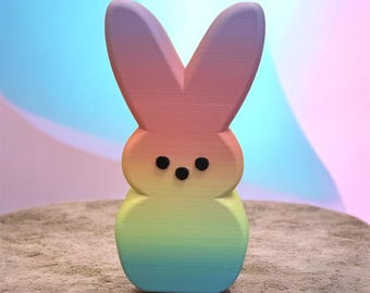 May include: A colorful, rainbow-hued bunny figurine with a gradient of colors from pink to blue. The bunny has black eyes and is standing on a textured surface. The background is a soft blend of blue and pink.