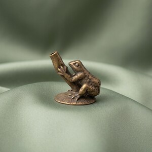 K&ouml;nnte beinhalten: Eine kleine, antik wirkende Metallfigur in Bronzet&ouml;nen, die einen Frosch auf einer strukturierten, kreisf&ouml;rmigen Basis zeigt. Der Frosch ist so dargestellt, dass er sich an einem hohlen, zylindrischen Rohr festh&auml;lt, das nach oben ragt.