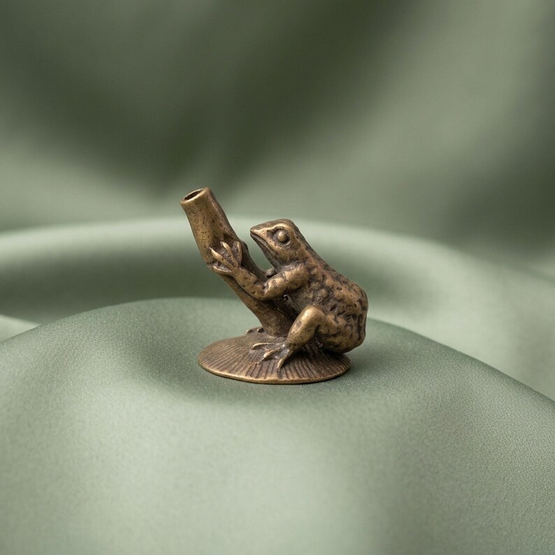 K&ouml;nnte beinhalten: Eine kleine, antik wirkende Metallfigur in Bronzet&ouml;nen, die einen Frosch auf einer strukturierten, kreisf&ouml;rmigen Basis zeigt. Der Frosch ist so dargestellt, dass er sich an einem hohlen, zylindrischen Rohr festh&auml;lt, das nach oben ragt.