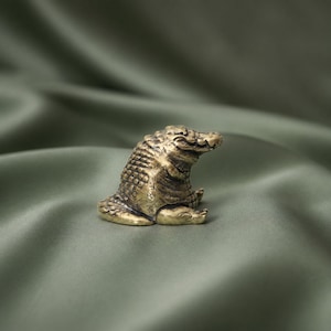 Könnte beinhalten: Eine kleine, metallische Messingfigur eines sitzenden Krokodils. Die Skulptur weist eine texturierte, schuppige Oberfläche mit einem dunklen, antik wirkenden Finish auf und zeigt das Tier in einer ruhenden Position mit leicht zur Seite gedrehtem Kopf.