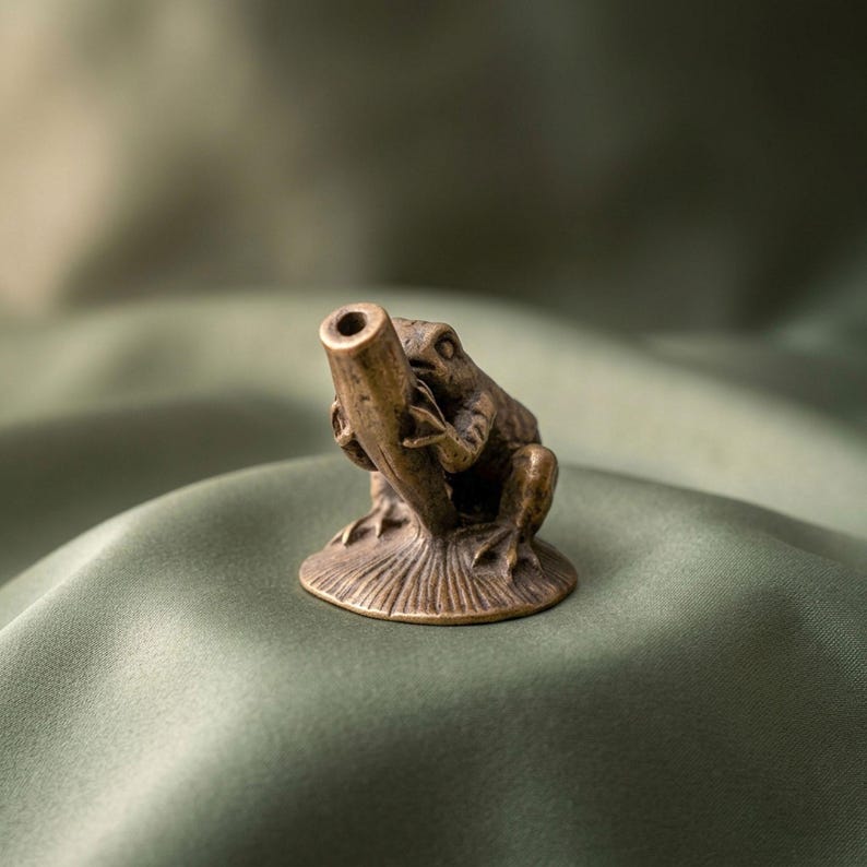 K&ouml;nnte beinhalten: Eine kleine bronzefarbene Metallfigur, die einen Frosch auf einem runden Sockel darstellt, der ein hohles, nach oben gerichtetes zylindrisches Rohr h&auml;lt. Das Objekt hat eine strukturierte Oberfl&auml;che mit detaillierten Gliedma&szlig;en und Augen.