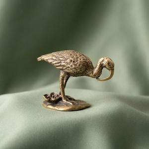 May include: A small brass figurine of a bird with a long, curved beak, standing on a textured base that includes a small flower. The metal has a weathered, antique finish with visible feather detailing across the body and wings of the bird.