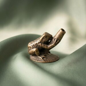 K&ouml;nnte beinhalten: Eine kleine, antik wirkende Messingfigur eines Frosches auf einem runden Sockel. Der Frosch sitzt in einer aufrechten Position und h&auml;lt ein hohles, zylindrisches, r&ouml;hrenartiges Objekt, das von seinen Vorderbeinen aus nach oben ragt.