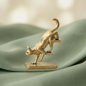 May include: A brass-colored metal figurine of a cat in a stretching pose, positioned on a rectangular base with two small steps. The cat is depicted with its front legs extended forward and its tail raised high in the air.